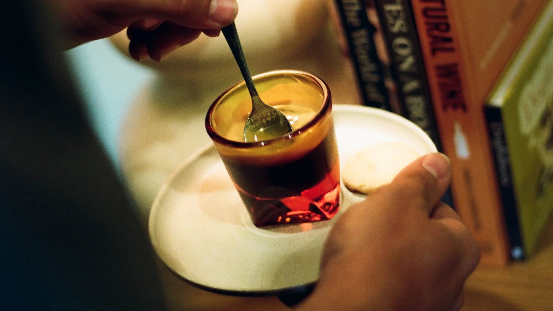 A hand stirring a dark coffee in an amber glass on a saucer beside a row of books