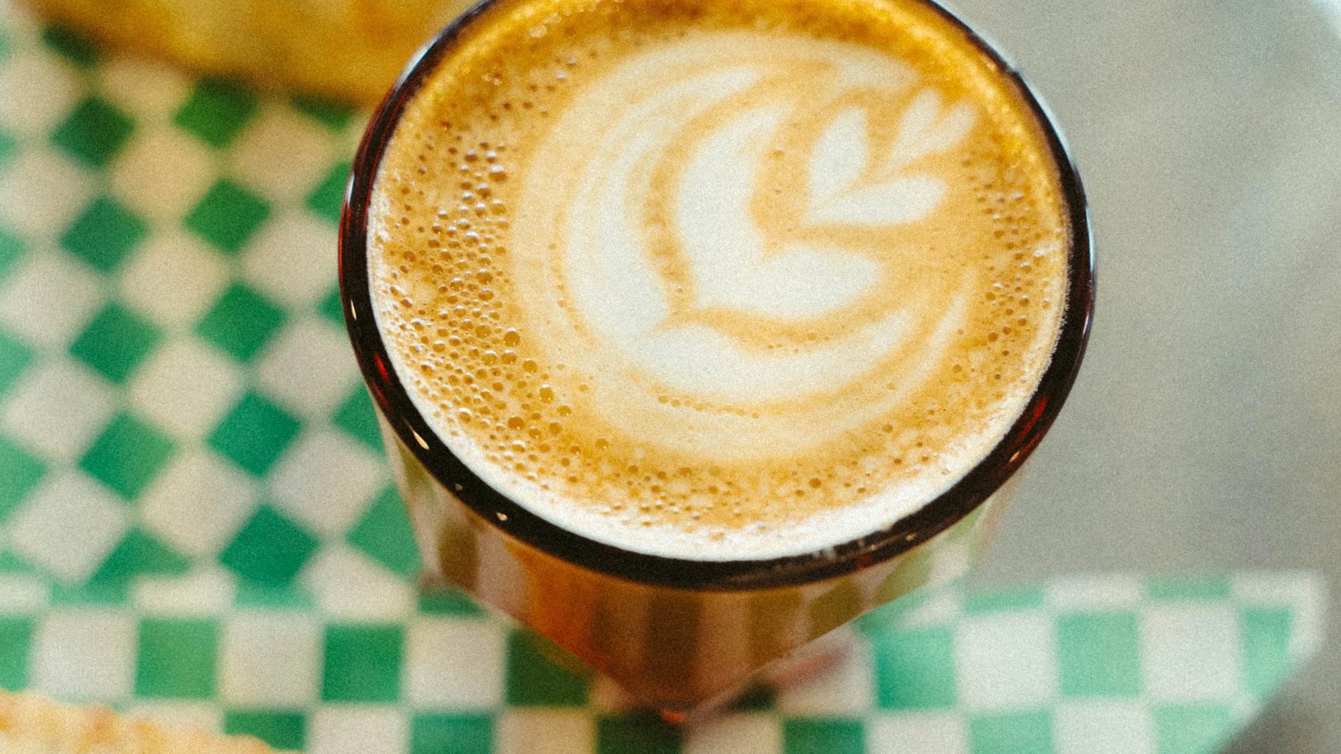 A latte with rosetta art in an amber glass beside pastries on green checkered paper at Abode Coffee